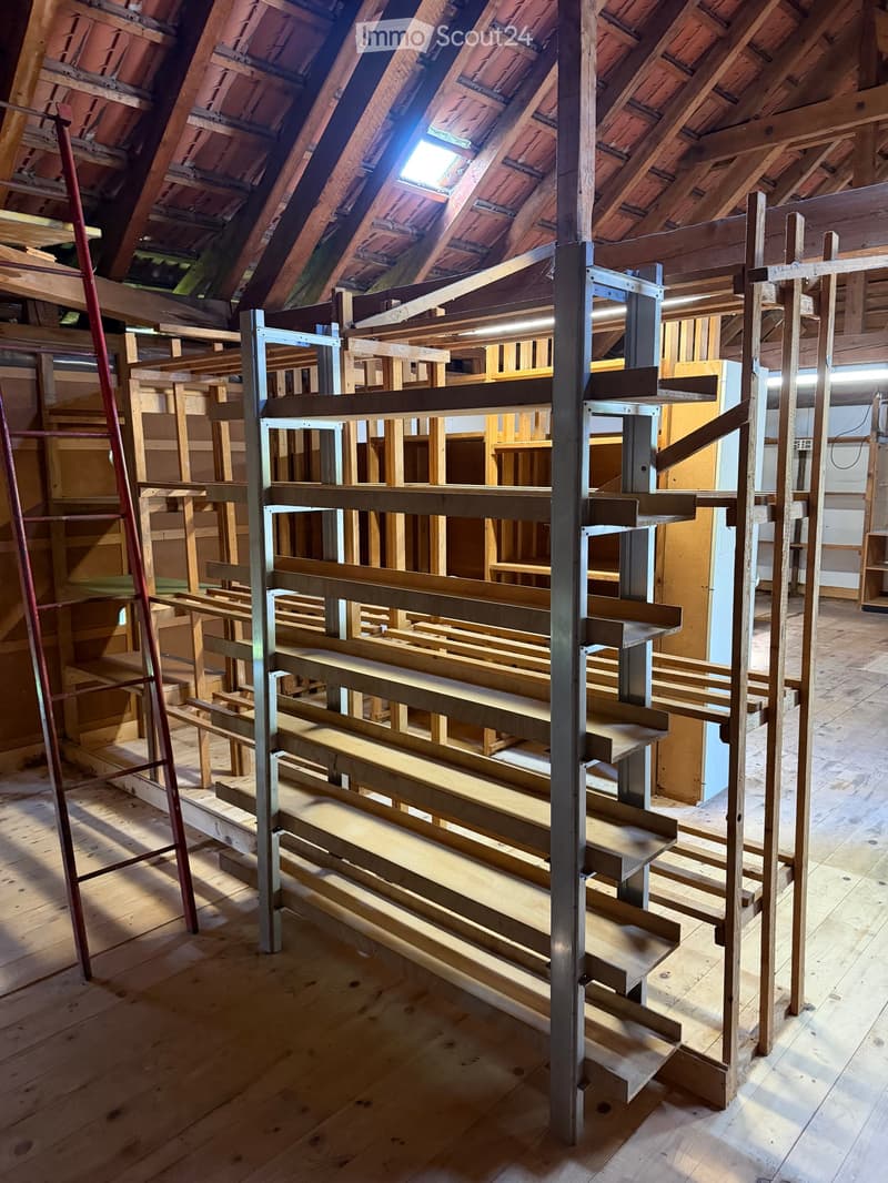 An attic space with wooden beams, exposed rafters, and a metal staircase frame. The floor is unfinished wood, and the ceiling has skylights allowing natural light to enter.