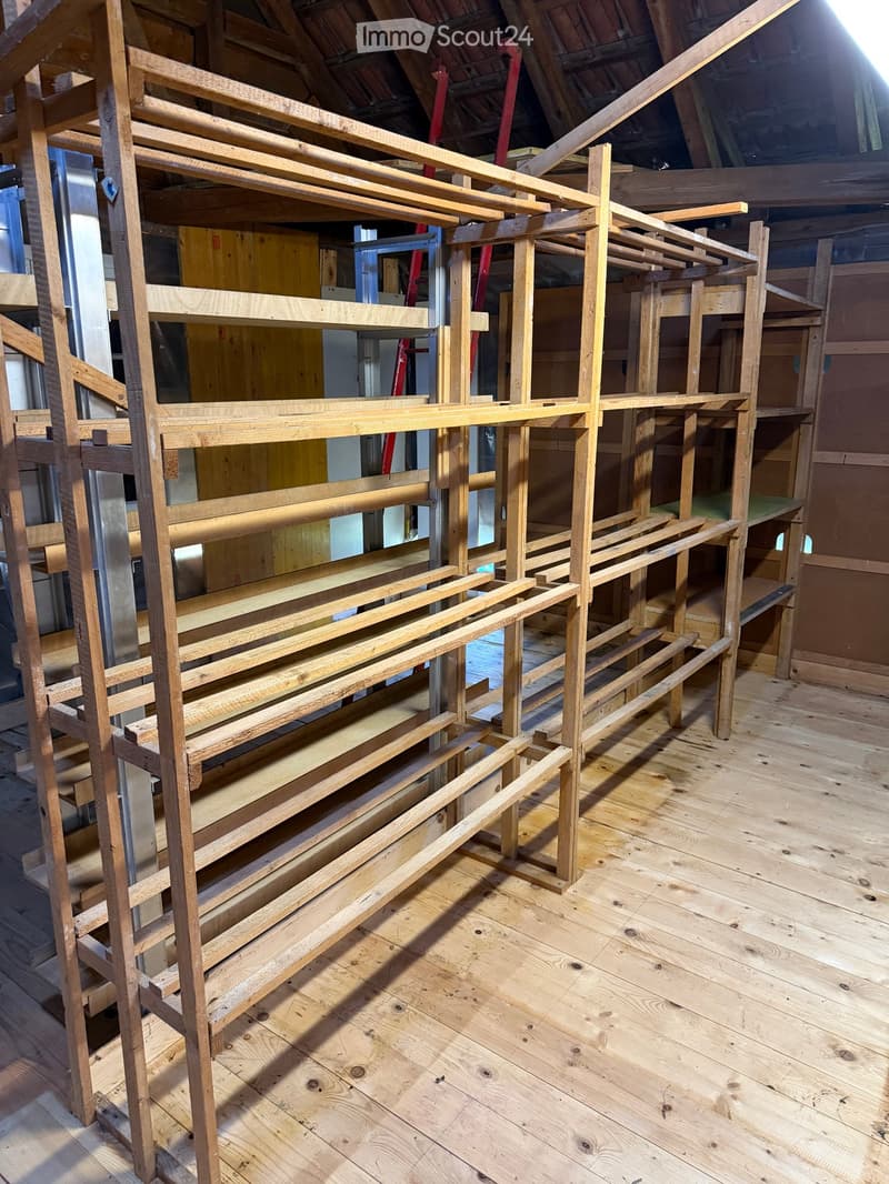 An unfurnished room with wooden flooring and a large wooden shelving unit under construction. The shelving unit features multiple levels and is supported by vertical wooden beams. The room has exposed wooden beams on the ceiling and walls.