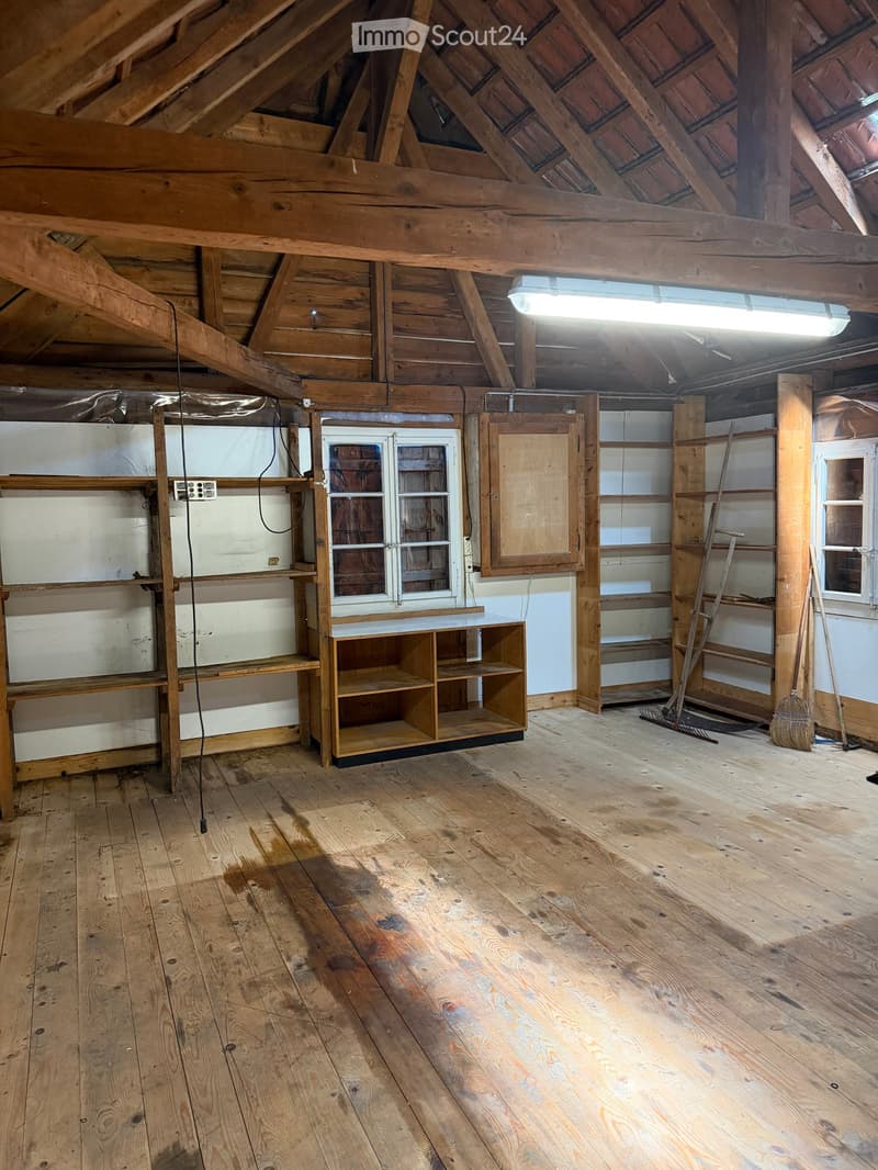 Empty attic space with wooden beams, exposed wooden floor, and minimal built-in shelving. No furniture or equipment present.