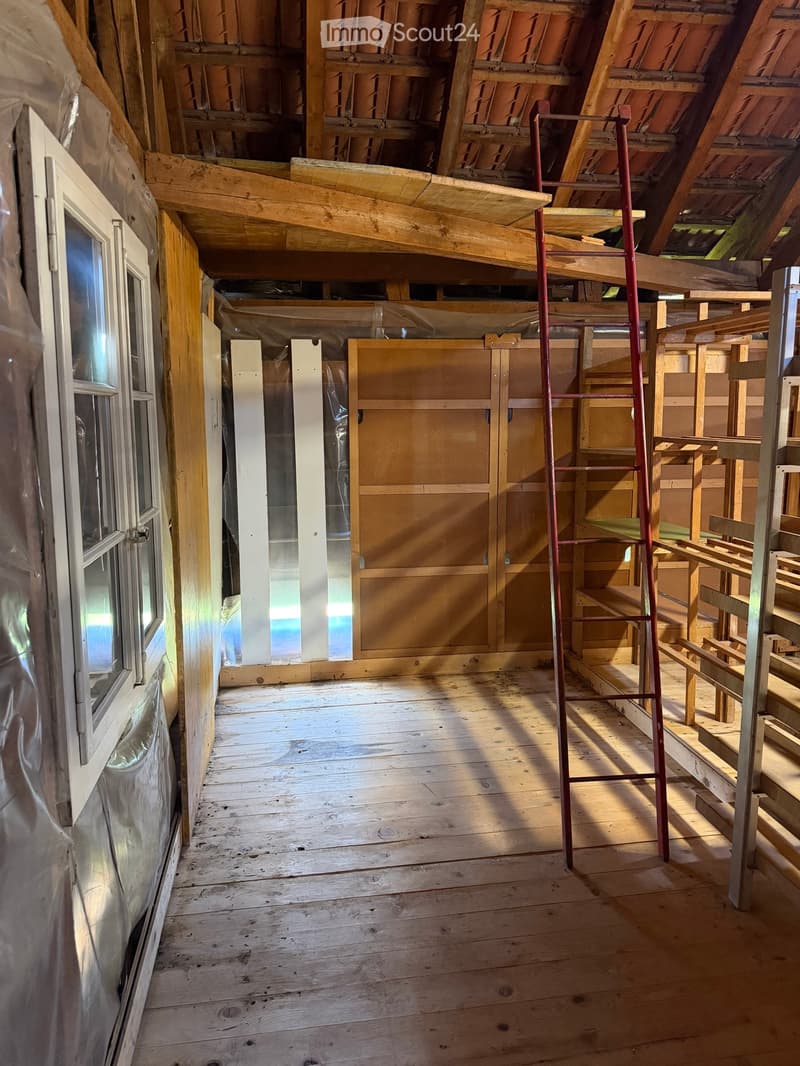 An empty attic space with wooden floors, exposed wooden beams, and a ladder leading to the roof. The room has a window on one side and is partially covered with plastic sheeting.