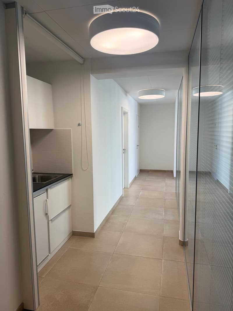 Modern hallway with white walls, beige tiled floor, and recessed lighting. Features a glass wall on one side and a small kitchenette area on the left.