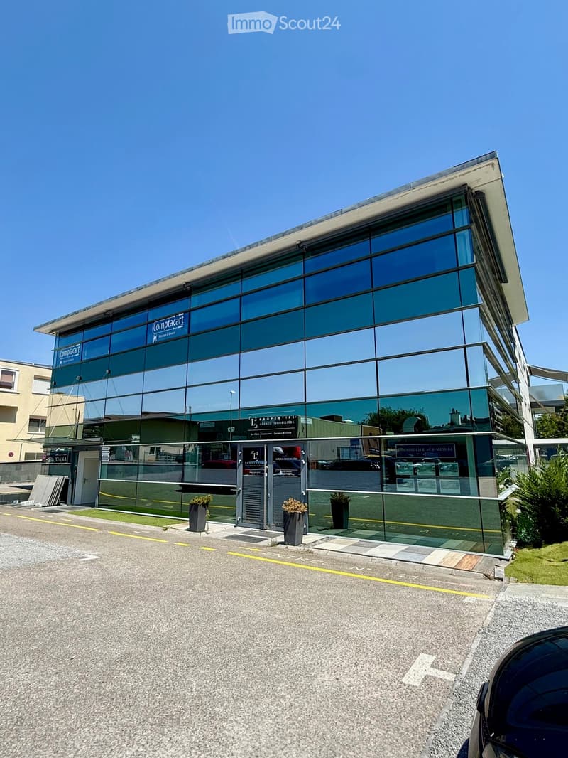 Modern commercial building with large glass windows and signage for 'Comptacart' and 'PROPERTIES' real estate agency. Features a clean, contemporary design with potted plants near the entrance.