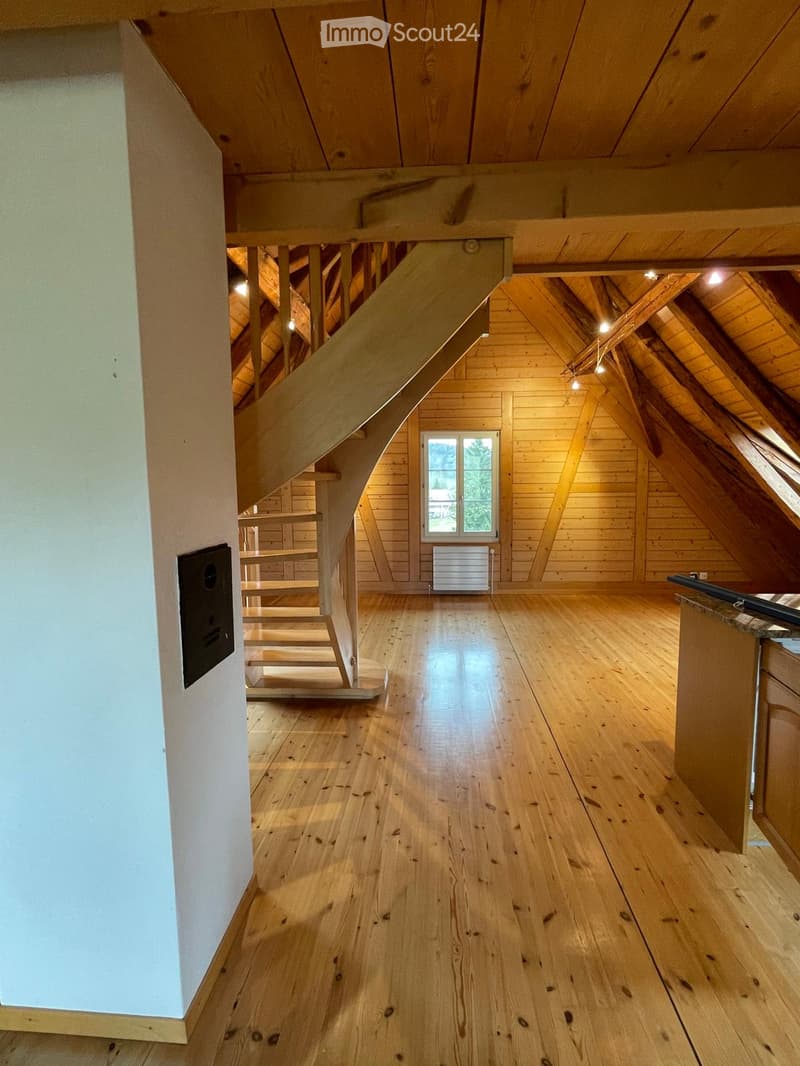 An empty attic space with wooden floors, exposed wooden beams, and a staircase leading to another level. The room features wooden walls and a window letting in natural light.