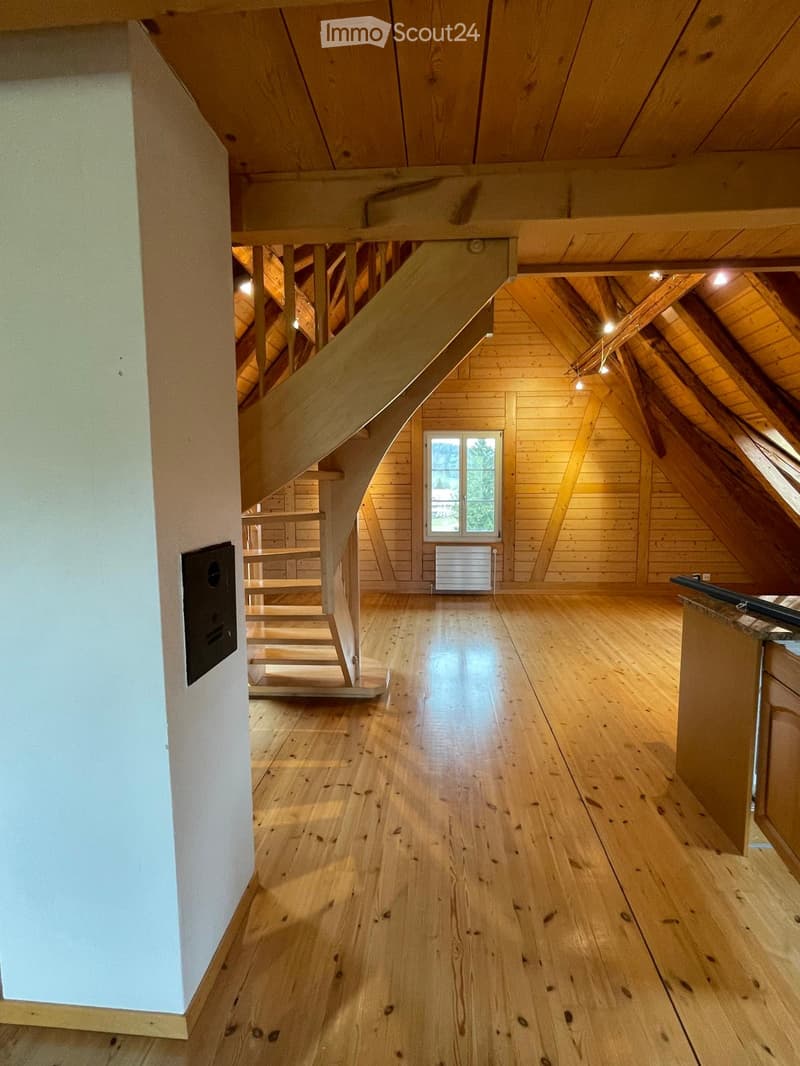 Empty attic space with wooden floors, exposed wooden beams, and a staircase leading to another level. There is a window with a view of greenery and a radiator below it.