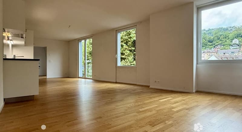 Empty room with wooden flooring, large windows, and a view of greenery and buildings outside.