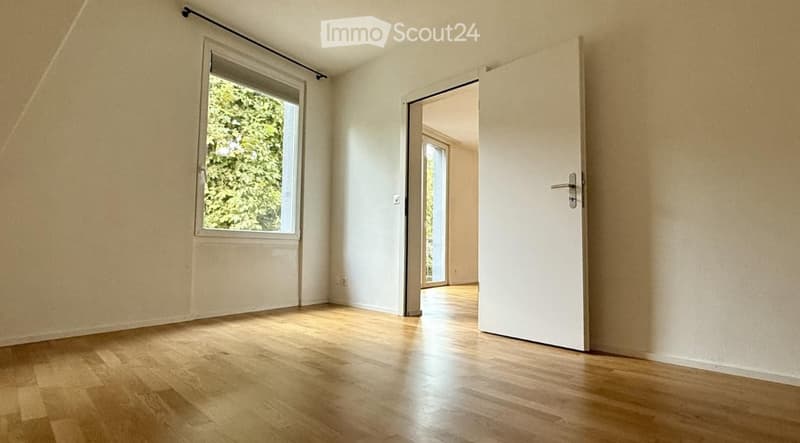 Empty room with wooden flooring, white walls, and a large window with a view of greenery. A white door is open, leading to another room with similar flooring and a large window.