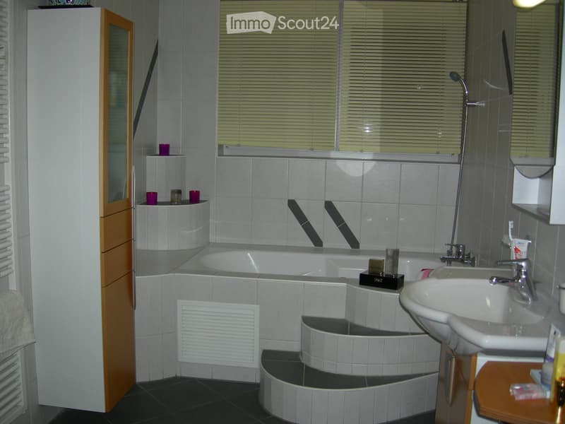 Modern bathroom with a bathtub, sink, mirror, and a small shelf with decorative items. The room features tiled walls and a window with blinds.