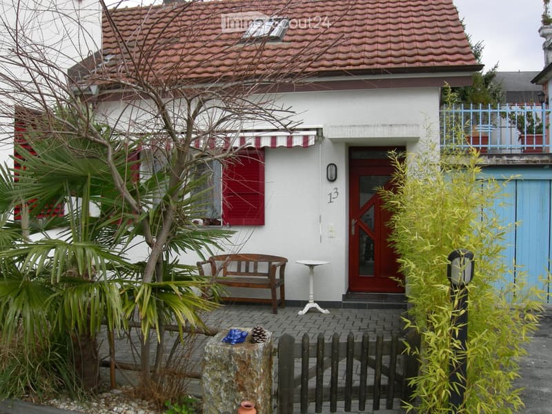 Two-story house with red-tiled roof, white walls, red shutters, and a small balcony with a railing. The entrance features a red door with a glass panel, and the house number "13" is displayed on the wall. The surrounding area includes a small garden with