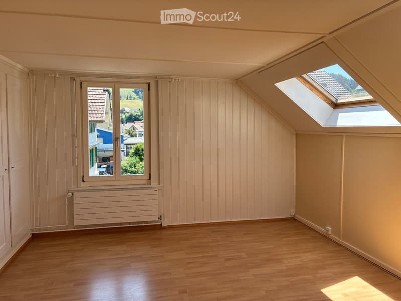 Empty attic room with wooden flooring, slanted ceilings, and skylights, featuring a window with a view of the neighborhood.