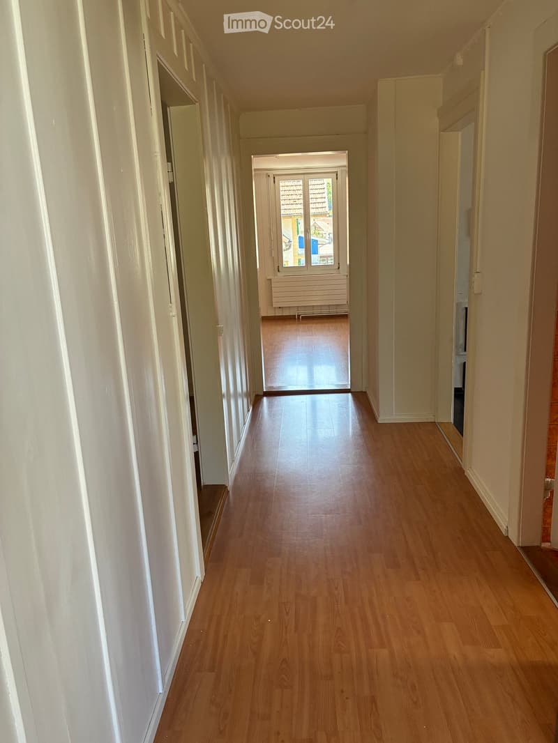 Empty hallway with wooden flooring, white walls, and a door leading to an outdoor area with a view of a tiled roof and surrounding buildings.