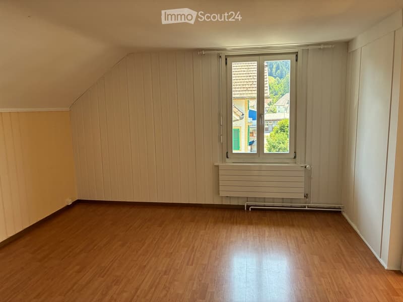 Empty room with wooden flooring, white walls, and a large window offering a view of the outside. The room features a radiator below the window and has a slanted ceiling on one side.