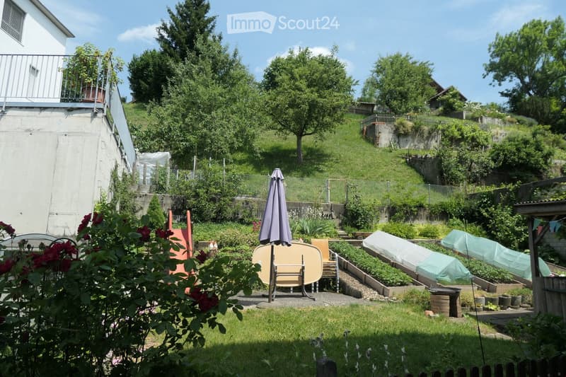 Garden with raised beds, patio furniture, umbrella, and surrounding greenery.