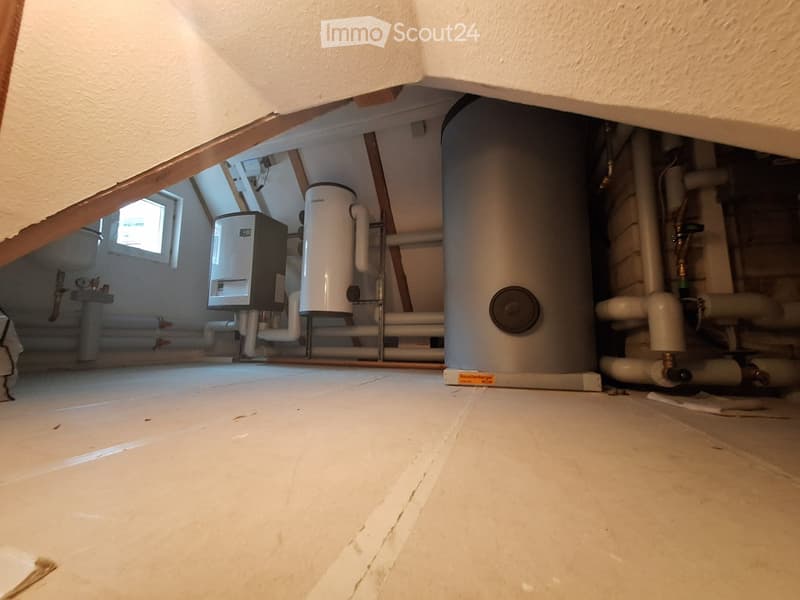 Empty basement space with exposed pipes, water heaters, and a small window, showing a concrete floor and wooden beams.