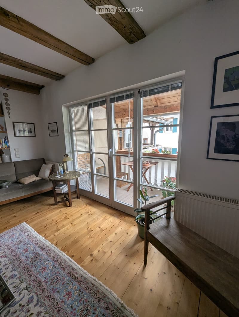 Cozy living room with wooden floors, large sliding glass doors leading to a balcony, a sofa, a small round table with a lamp, and a potted plant near the entrance.