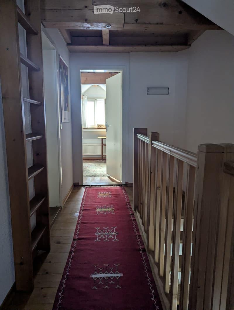 A narrow hallway with wooden floors, a red runner rug, and a wooden staircase railing on the right side. An open doorway leads to another room with a small table and chair visible.