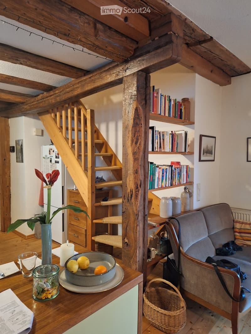 Cozy living room with wooden beams, a staircase, a bookshelf filled with books, a sofa, a small table with a bowl of fruit, a vase with a plant, and a woven basket on the floor.