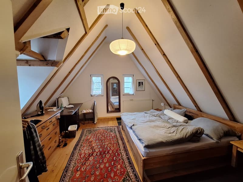 Cozy attic bedroom with wooden beams, slanted ceilings, and large windows. Features a double bed, wooden desk, mirror, and a patterned rug.