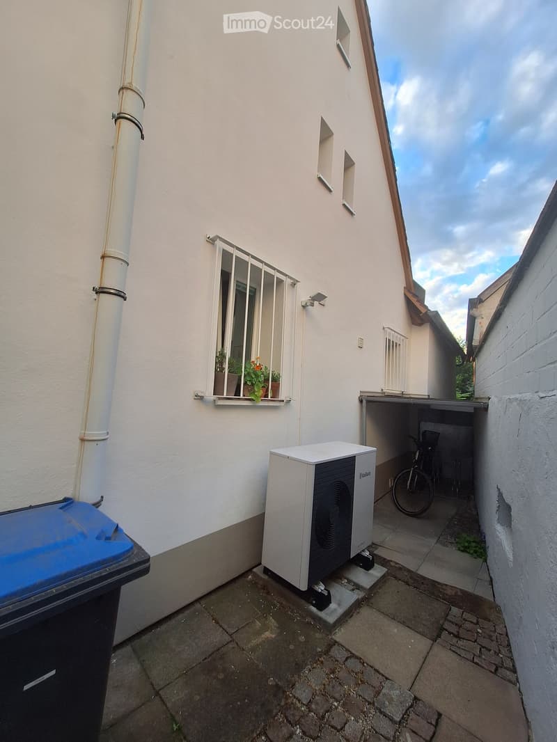 Narrow outdoor area with a white wall, small window with potted plants, air conditioning unit, bicycle, and a blue trash bin.
