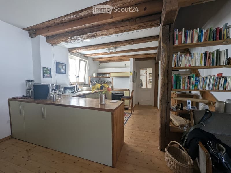 Open-plan living and kitchen area with wooden beams, kitchen island, bookshelf, and various appliances.