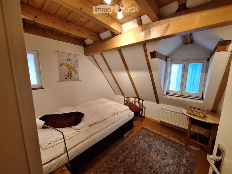 Cozy attic bedroom with wooden beams, slanted ceilings, and large windows. Features a single bed, a small wooden chair, a table with a basket, and a patterned rug.