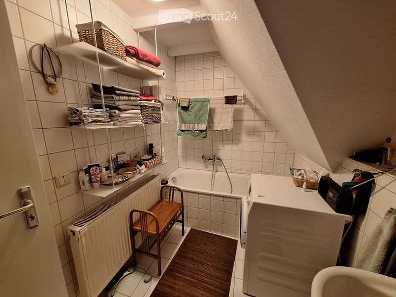 Bathroom with a bathtub, sink, wooden bench, shelves holding towels and baskets, and a radiator. The room features white tiled walls and a slanted ceiling typical of an attic space.