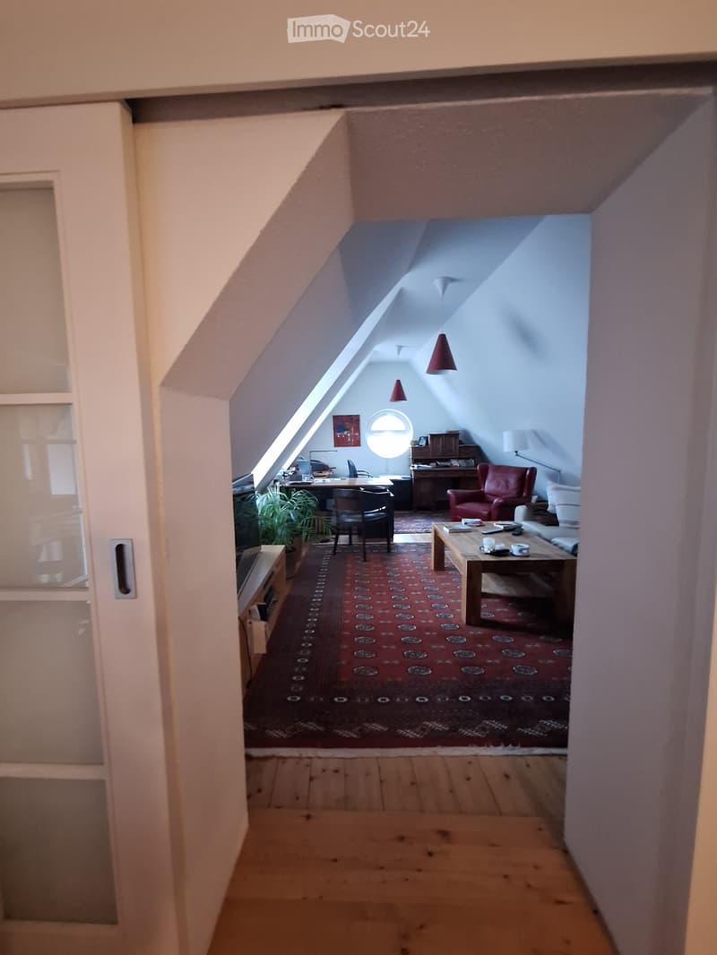 A spacious living room with wooden floors, a red patterned rug, a sofa, coffee table, chairs, desk, and a plant. The room features large windows and a slanted ceiling with skylights.