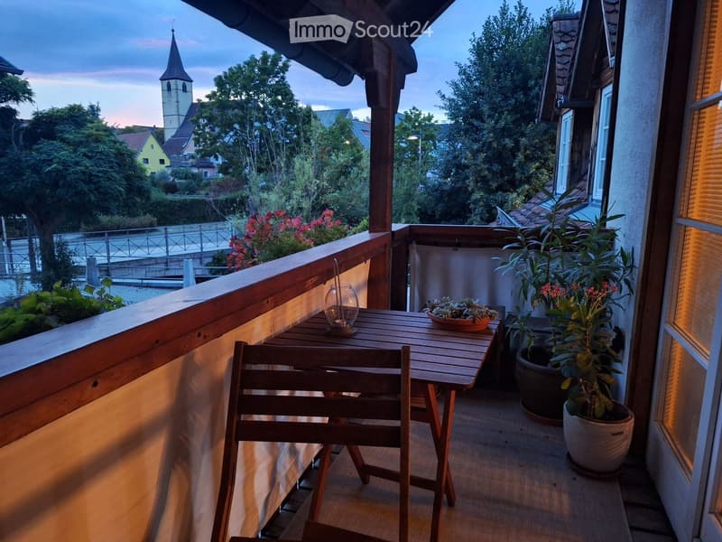 Balcony with wooden table, chairs, potted plants, and a view of a town with a church tower.