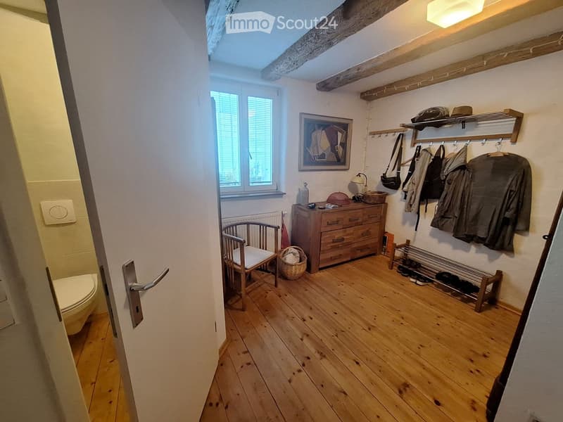 Cozy hallway with wooden flooring, a small chair, a drawer unit, coat rack, shoe rack, and a basket. A framed picture hangs on the wall, and a toilet is visible through an open door.
