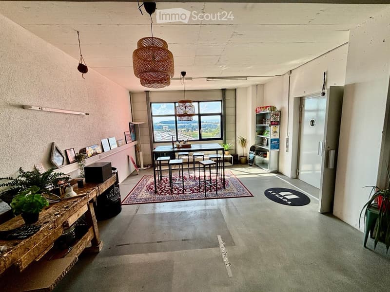 Open-plan space with a dining area, bar counter, and shelves. Features a large window with a scenic view, wicker pendant lights, and potted plants.