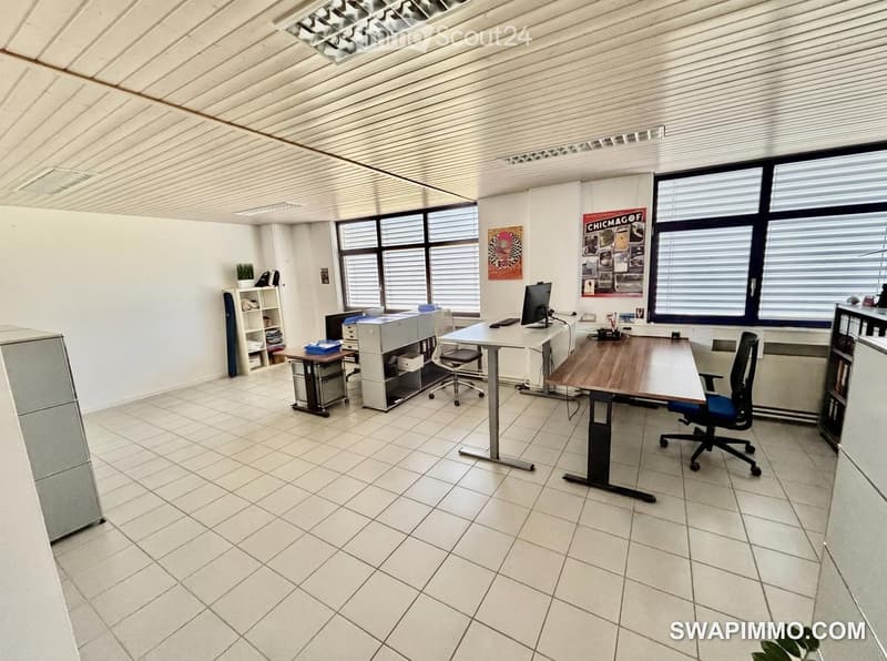 Office space with tiled floor, large windows, desks, chairs, a screen, printer, and shelves.