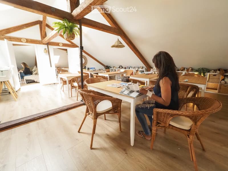Bright, airy room with wooden beams and exposed ceiling, featuring a mix of wicker and wooden furniture, including tables, chairs, and a sofa, with shelves and potted plants adding a cozy touch.