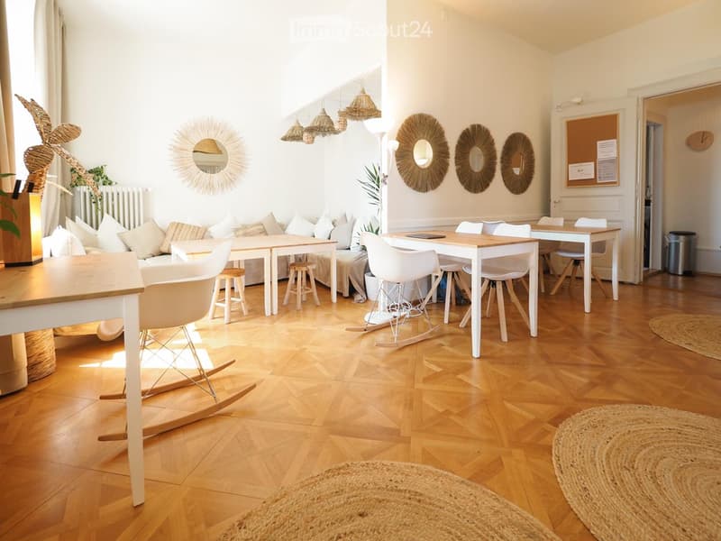 Bright, open living space with wooden flooring, white walls, and modern decor. Features a sofa with cushions, coffee table, dining table with chairs, rocking chair, and decorative mirrors on the wall.