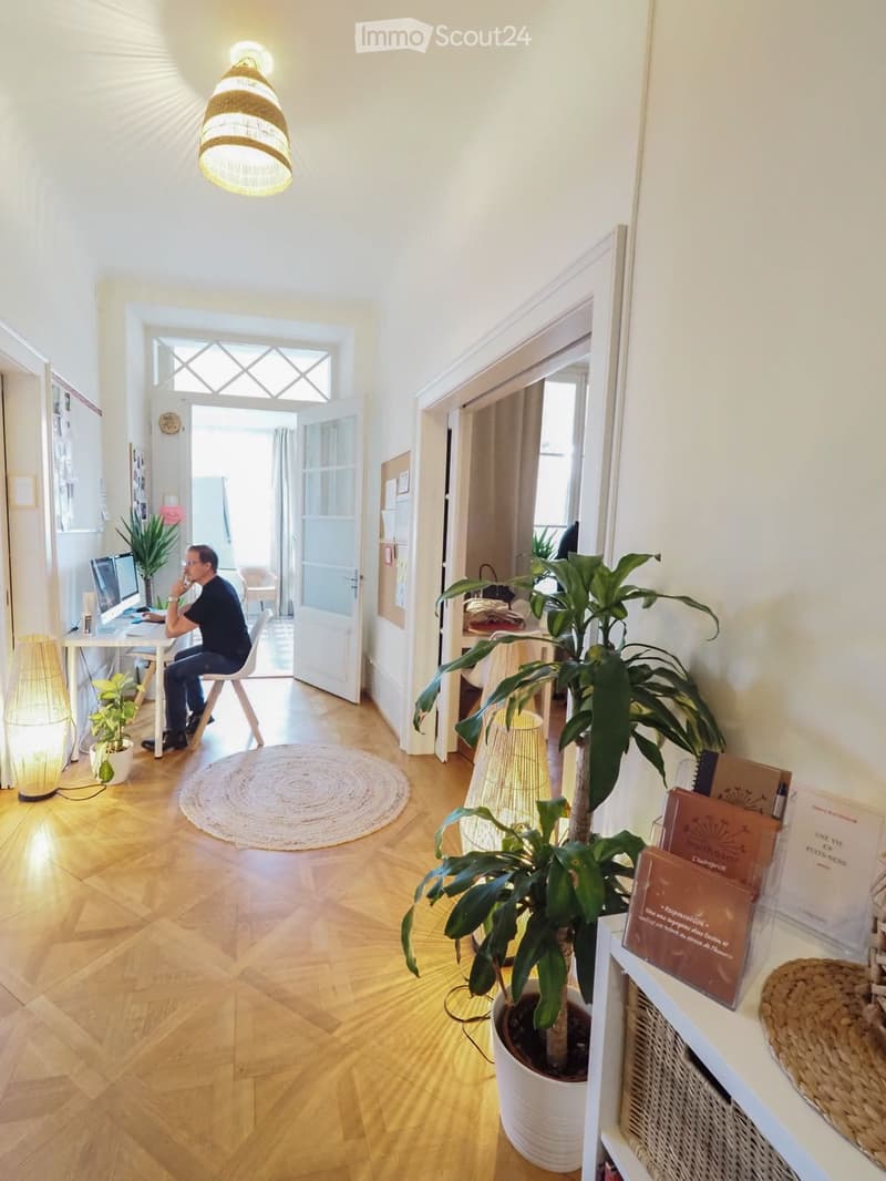 Bright, open living space with wooden flooring, a round woven rug, a hanging lamp, a desk with a computer, and several potted plants.