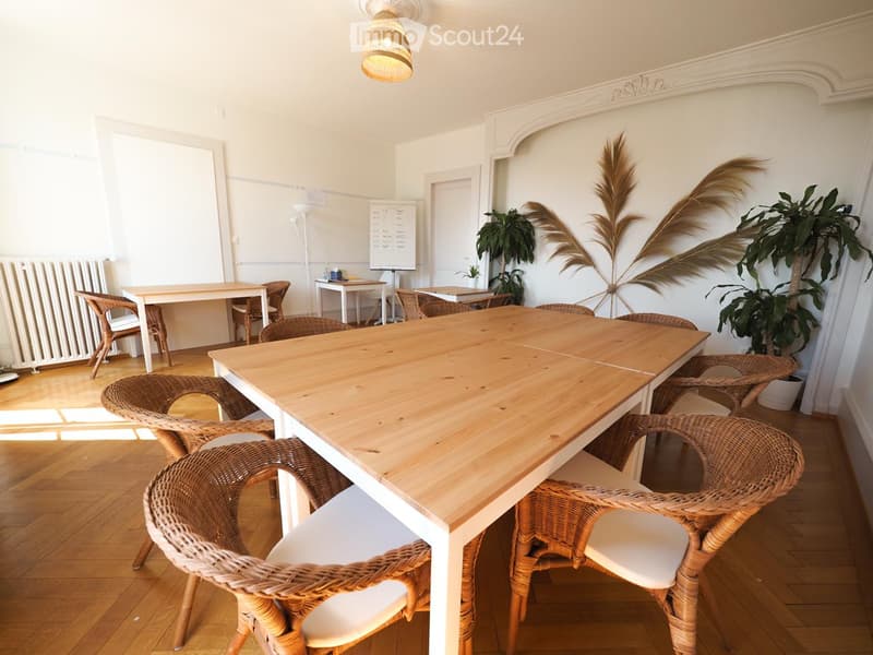 Bright room with wooden floor, large central table surrounded by wicker chairs, radiator, screen, and decorative plants.