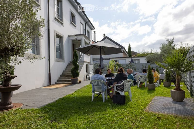 Garden area with a white building in the background, featuring a patio with a table and chairs, an umbrella, potted plants, and a small staircase leading to the building entrance.