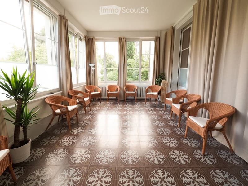 Bright balcony with large windows, wicker chairs with white cushions, patterned tile floor, and potted plant.