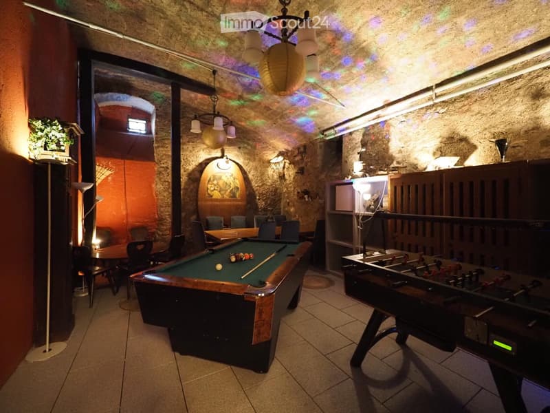 A cozy game room with a pool table, foosball table, and seating area. Exposed stone walls and warm lighting create a relaxed atmosphere.