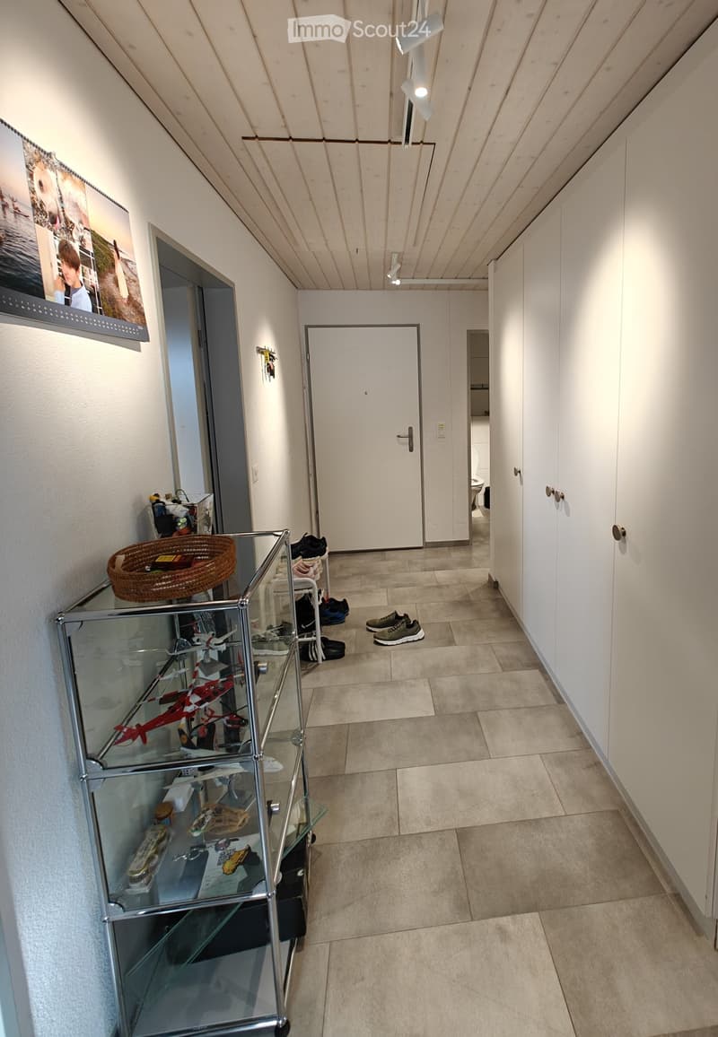 A modern hallway with tiled flooring, white walls, built-in wardrobes, and a glass display shelf holding toys and shoes. A framed picture hangs on the wall, and a door leads to another room.