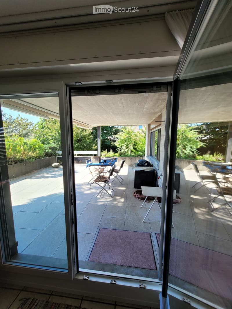 Balcony with tiled floor, glass sliding doors, outdoor furniture including a table, chairs, a grill, and a small stool. The balcony has a view of a garden with trees.