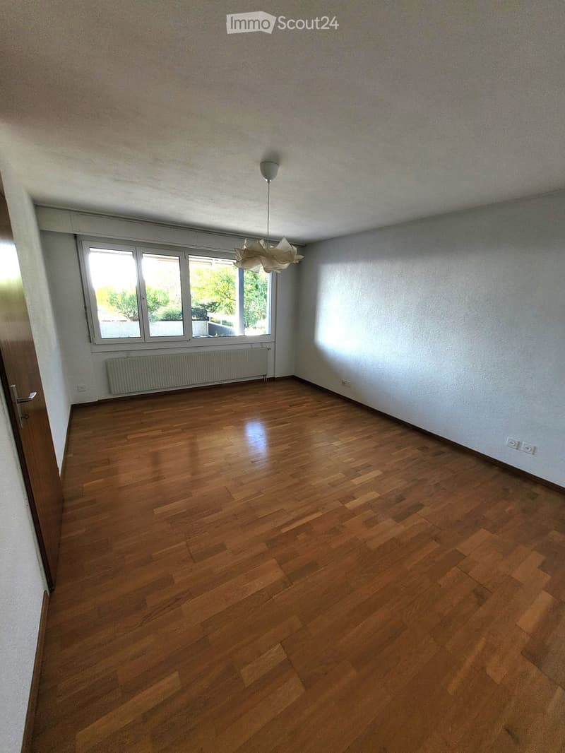 Empty room with wooden flooring, white walls, large window with blinds, radiator, and a hanging light fixture.