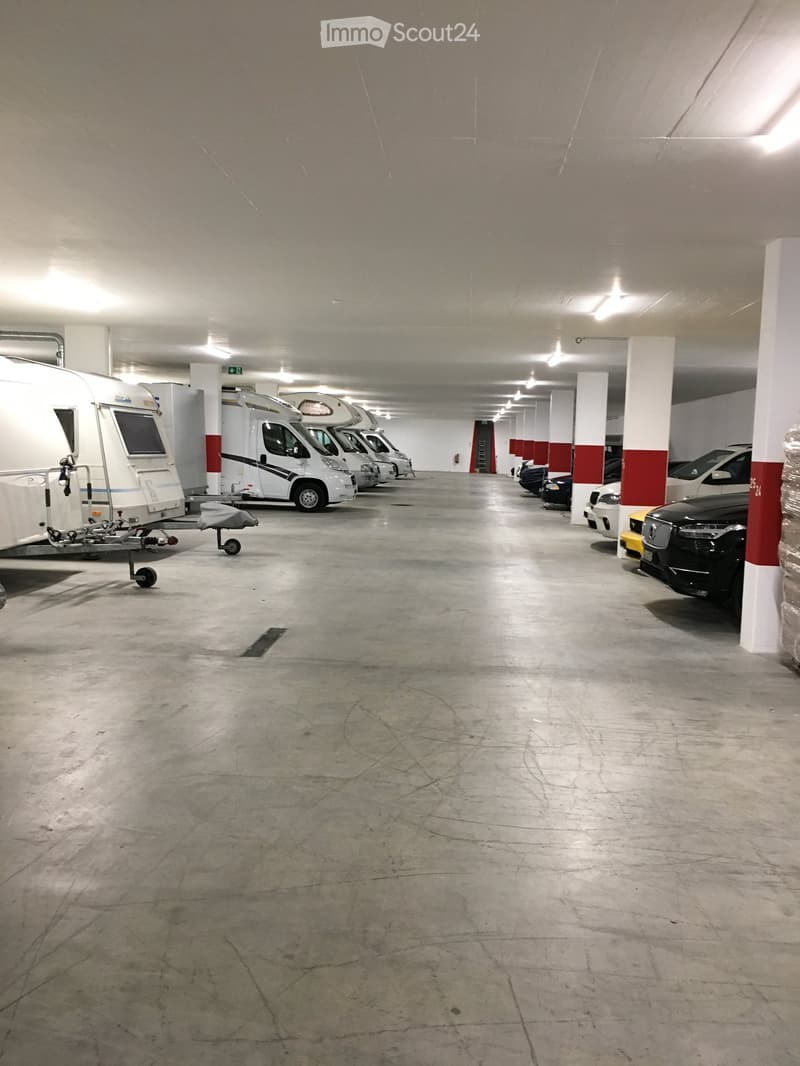 Underground parking area with several parked vehicles including cars and a camper van, concrete floor, white and red pillars, and overhead lighting.
