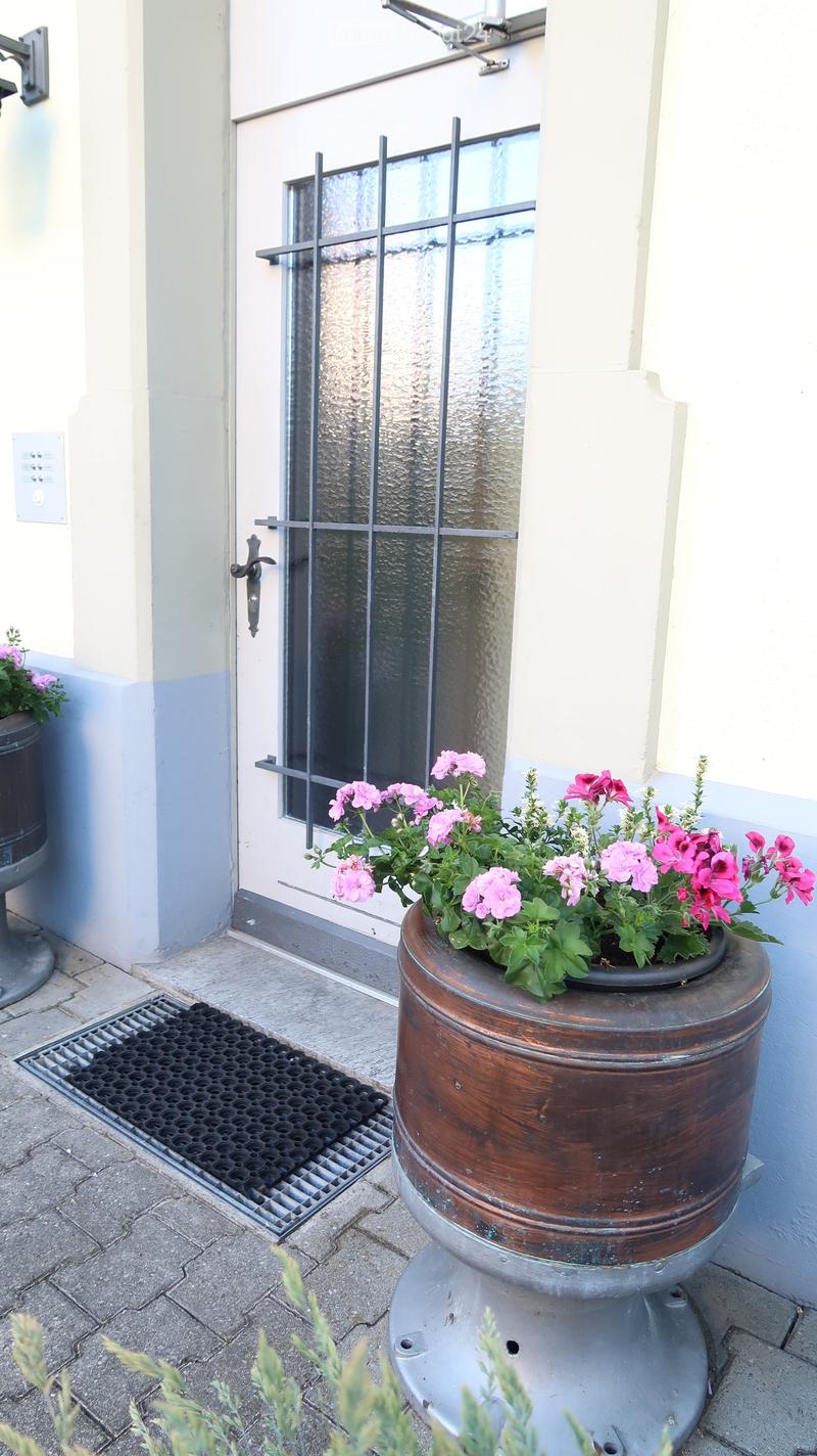 A decorative planter with pink flowers sits on a stone platform near a glass door entrance. The door features a metal handle and a small mat in front.