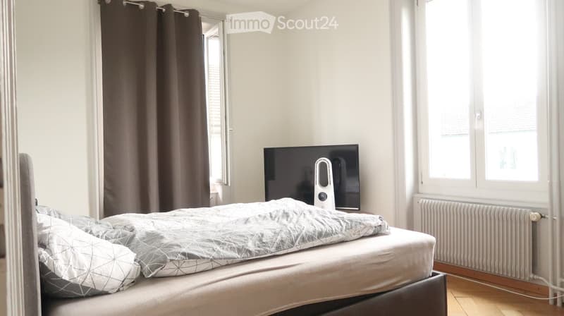 A modern bedroom with a neatly made bed, a flat-screen TV, and large windows with curtains. The room features light-colored walls and wooden flooring.