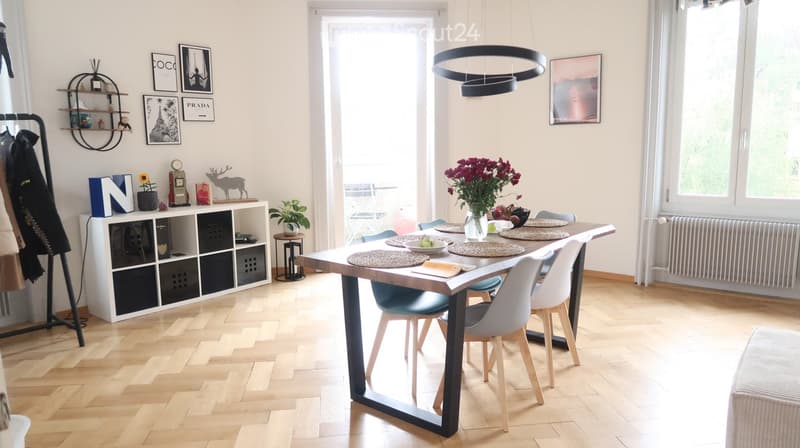 Dining room with wooden floor, large windows, modern dining table with chairs, flower vase, shelves with decorative items, and picture frames on the wall.
