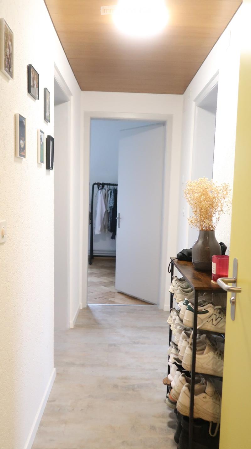 A narrow hallway with light-colored walls, a wooden ceiling, and a tiled floor. A shoe rack with multiple pairs of shoes is visible on the right, along with a small table holding a vase with dried flowers and a red cup. An open door leads to another room