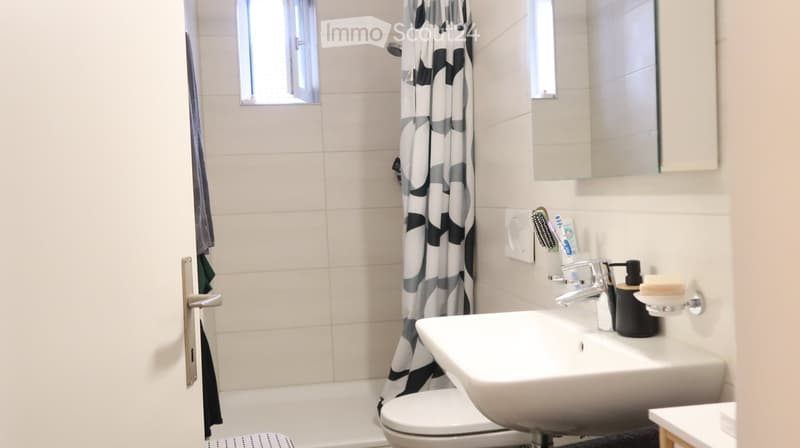 Modern bathroom with white tiles, a sink, toilet, bathtub, and a shower curtain. A mirror is mounted above the sink, and personal care items are placed on the sink counter.