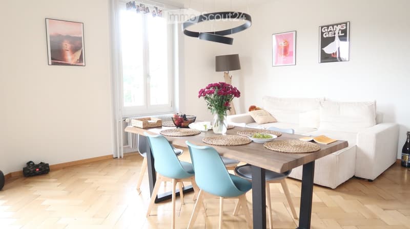 Bright living room with a white sofa, wooden dining table, modern chairs, and decorative items including a vase with flowers and picture frames on the wall.