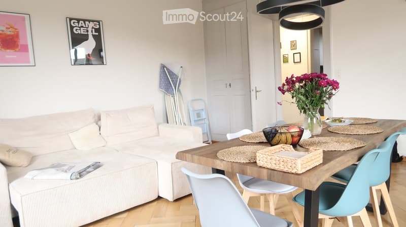 A modern living room with a white sofa, a wooden coffee table, colorful chairs, and a vase with flowers on the dining table.