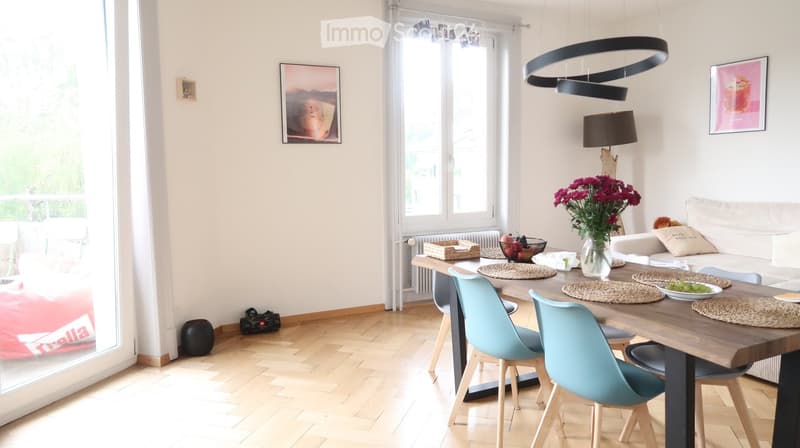 Bright living and dining area with wooden flooring, large windows, a white sofa, a wooden dining table with four chairs, a vase with flowers, and decorative items.