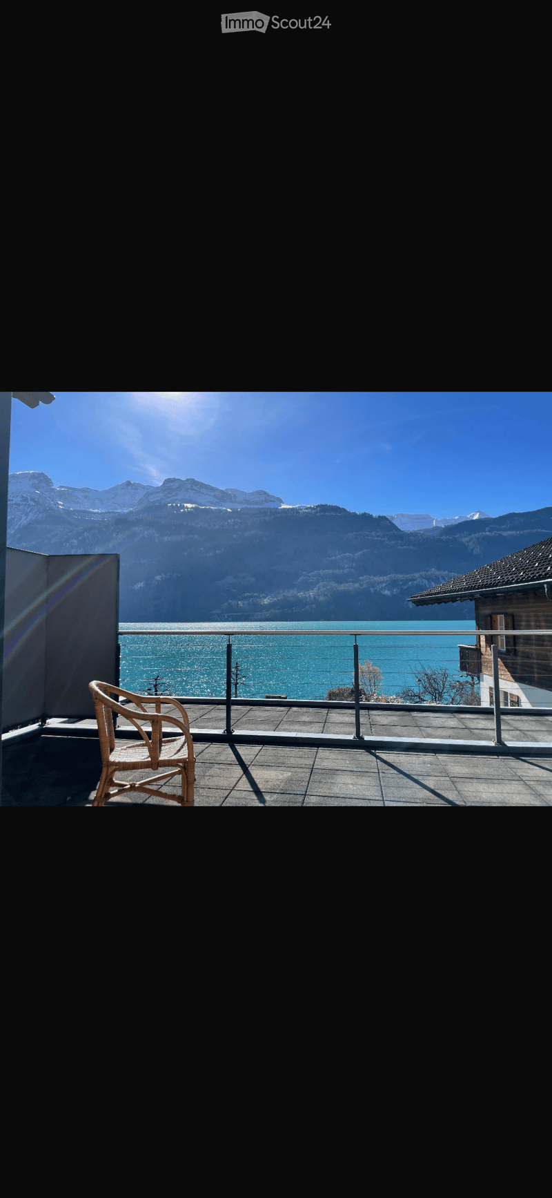 Balcony with a view of a lake and mountains, featuring a single wicker chair and a metal railing.